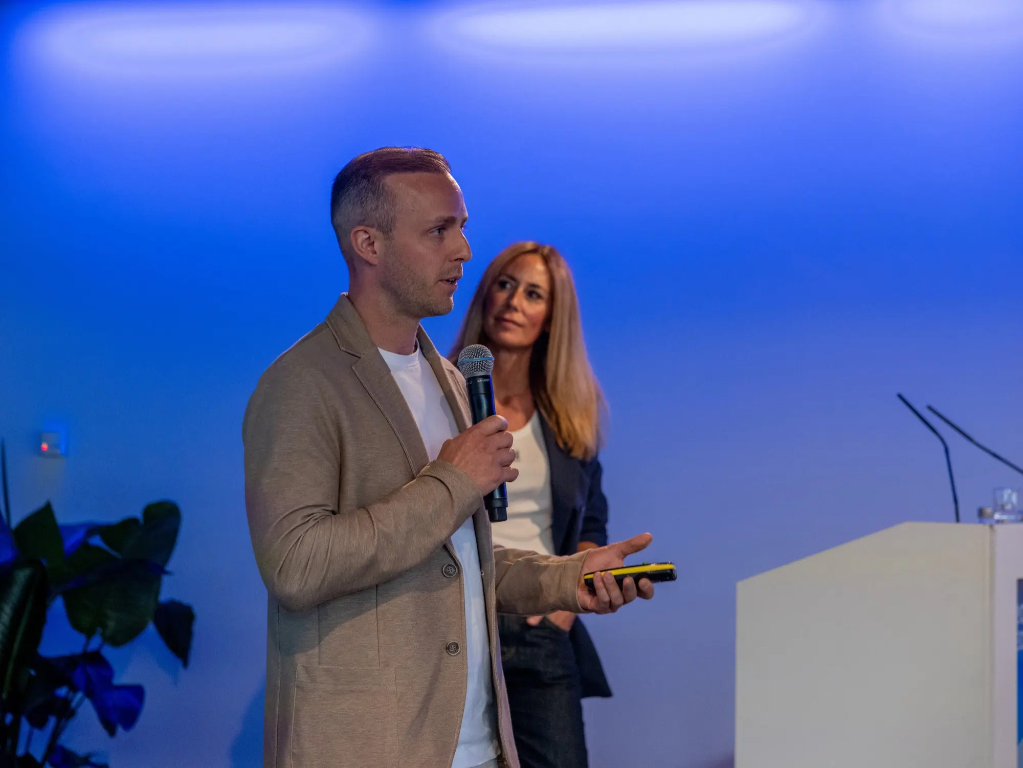 Dom Carter speaking on stage with a handheld microphone, a co-presenter beside him